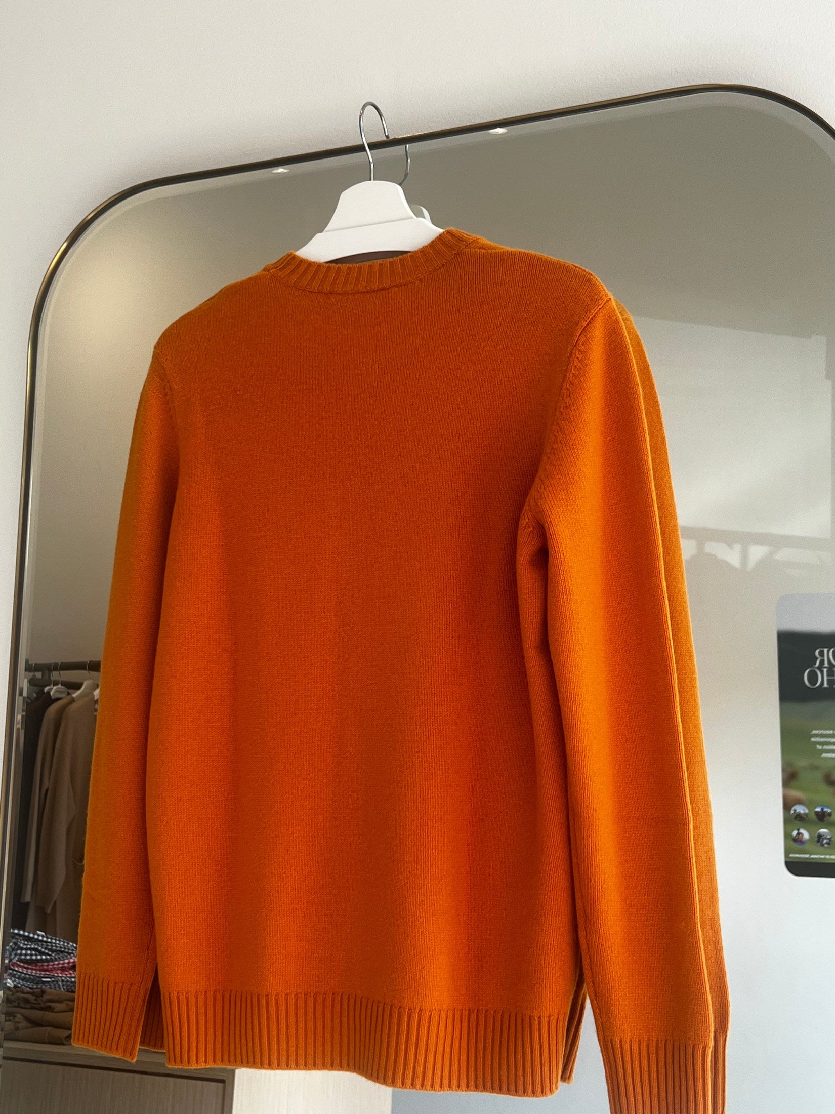 Orange round neck cashmere sweater hanging on white hanger in front of mirror for women’s fashion.