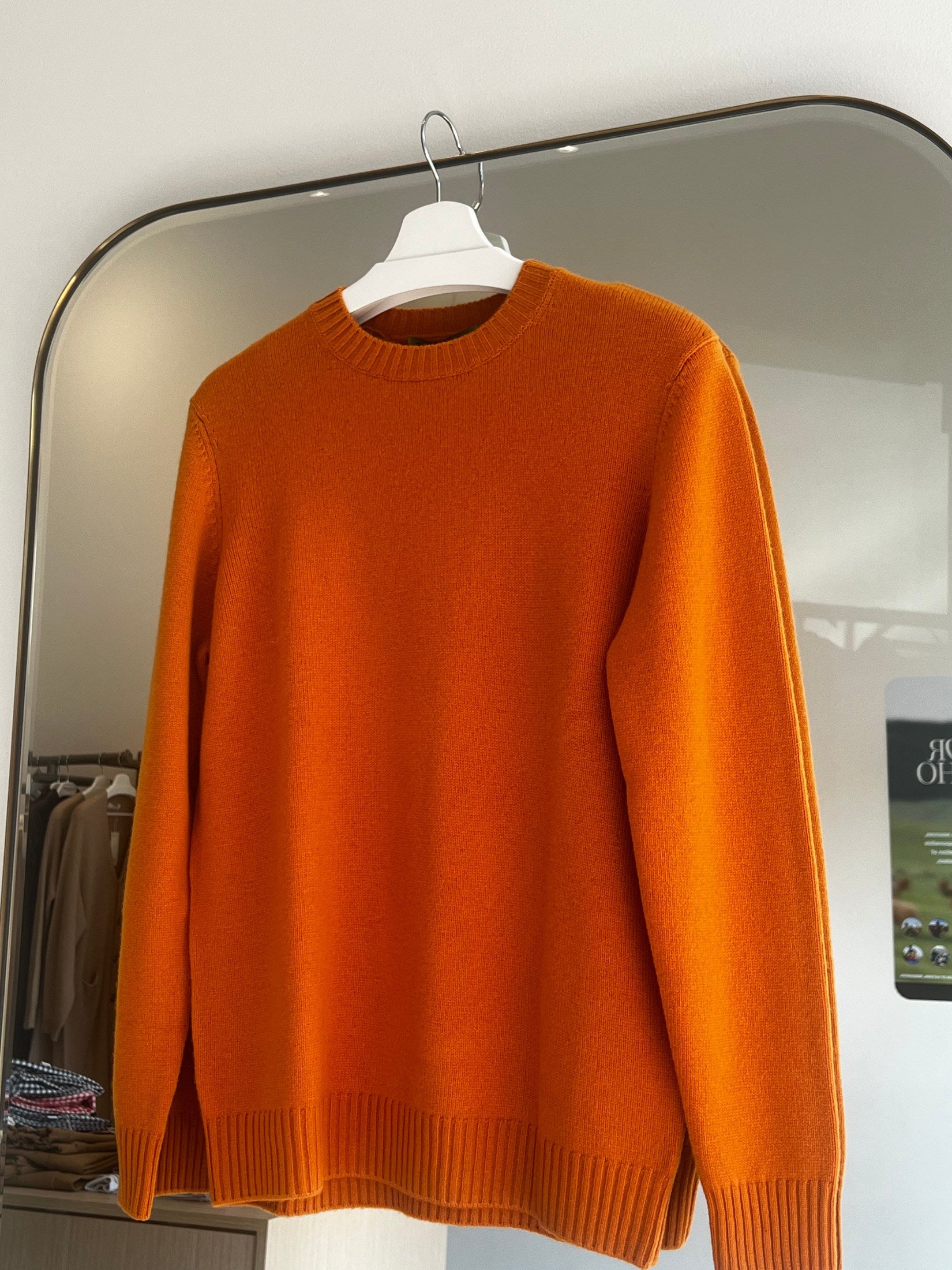 Orange round neck cashmere sweater hanging on white hanger with ribbed cuffs and hem in store setting