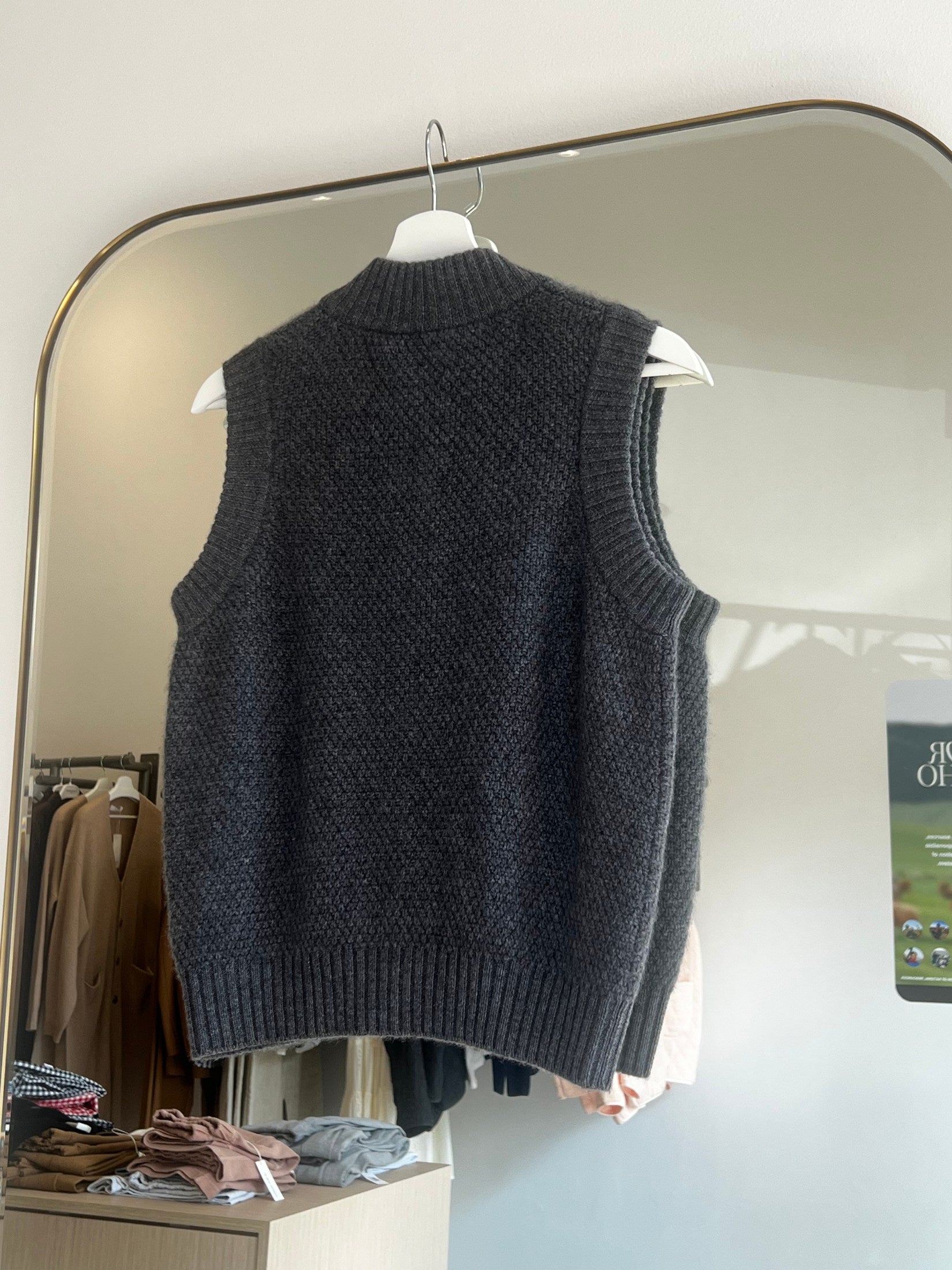 Dark gray moss stitch cashmere button vest hanging on a white hanger in front of a mirror in a boutique.
