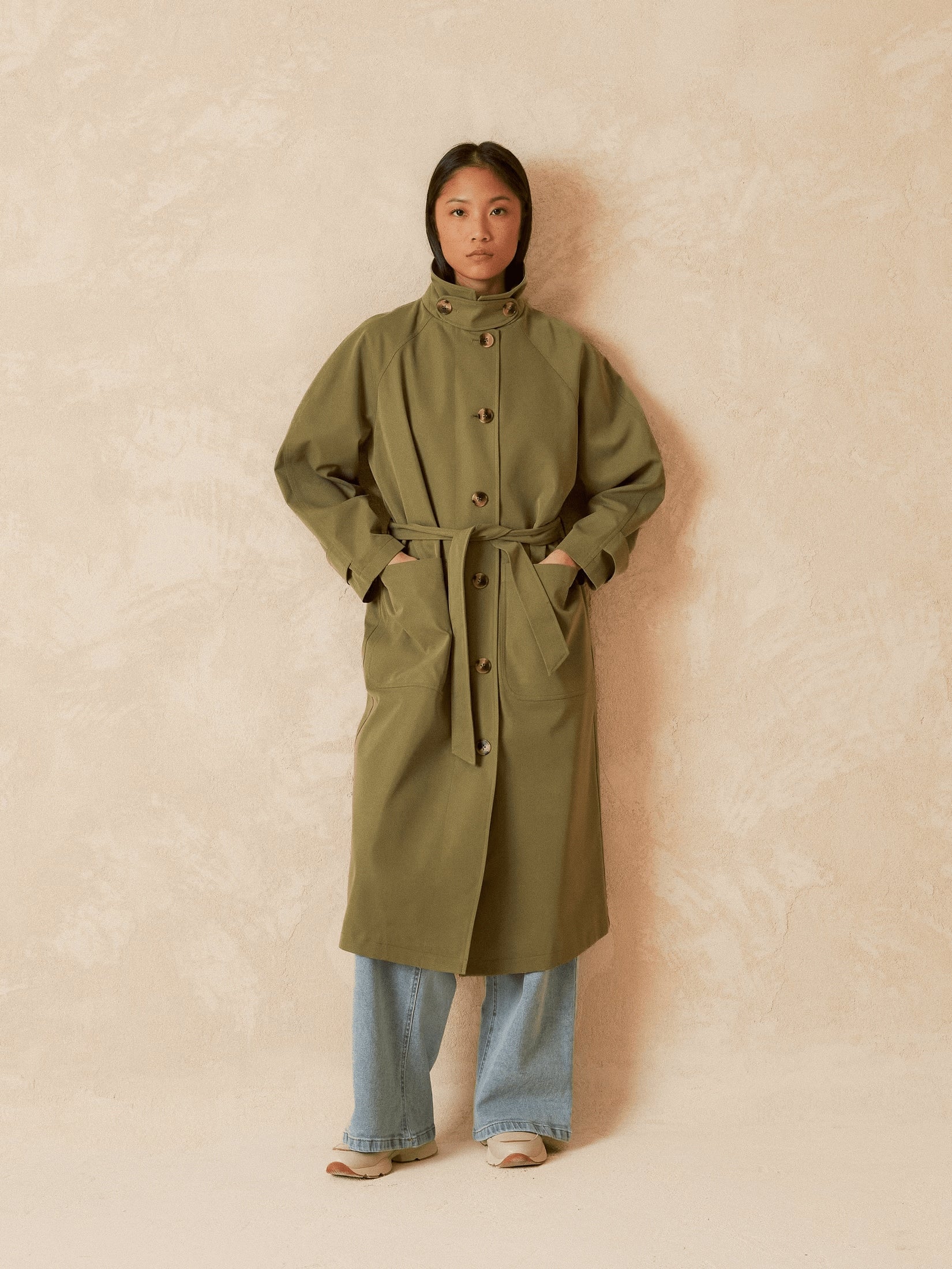 Front view of a model wearing a long khaki trench coat with belt.